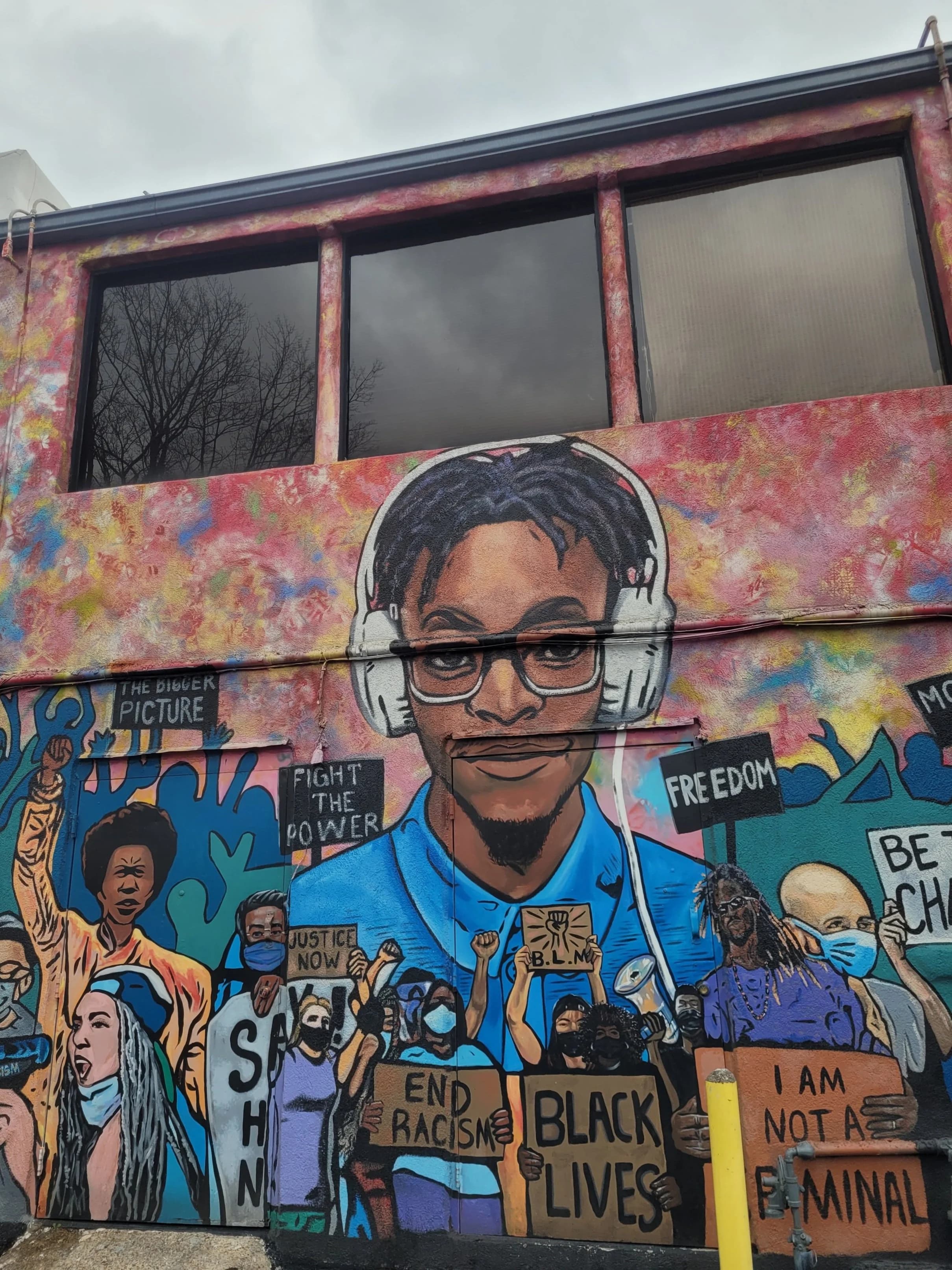 Street mural in Norfolk of a young Black man in headphones above a crowd holding signs for justice and Black lives