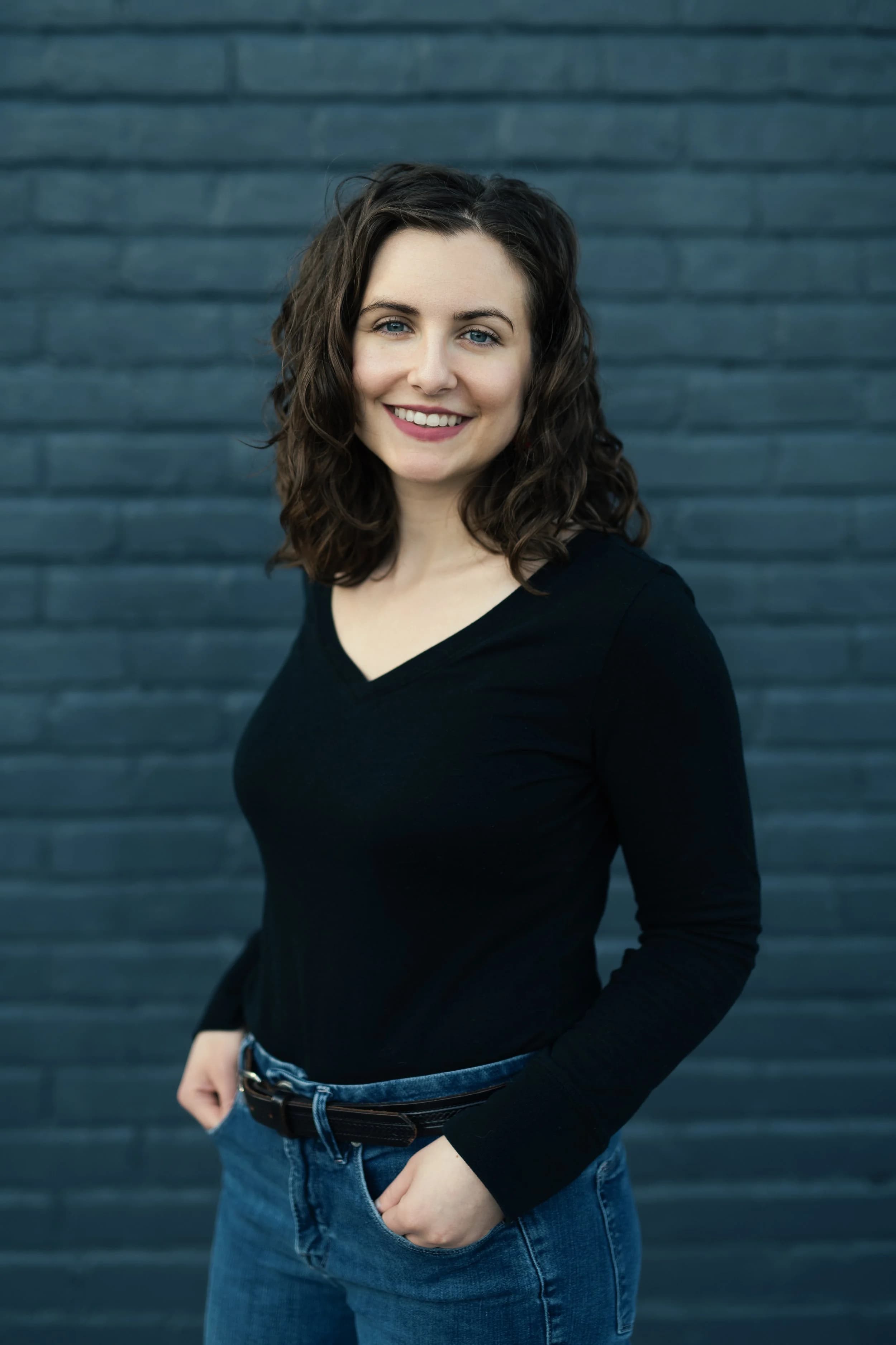 Dr. Renae DeLucia in a black top, smiling, in front of a blue-gray brick wall