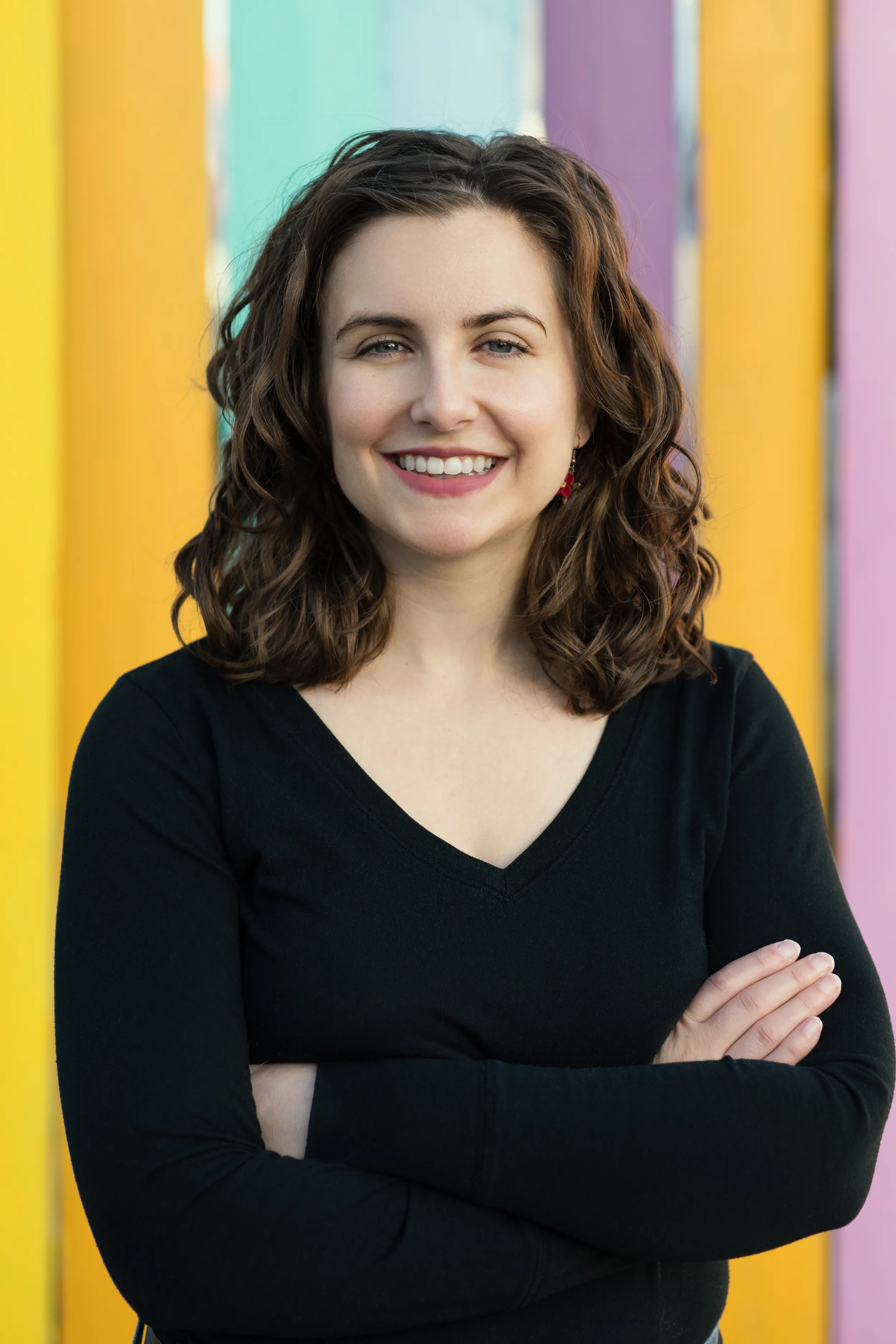 Dr. Renae DeLucia smiling with her arms crossed in front of a wall painted in yellow, teal, purple, and pink stripes
